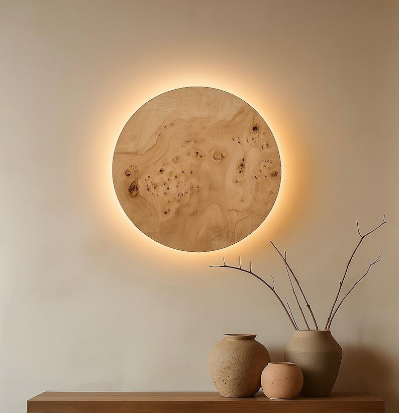 May include: A round, wooden wall light fixture with a warm, glowing halo. The wood grain is visible, and the fixture is mounted on a neutral-colored wall. Decorative vases and branches are in the foreground.