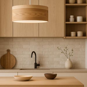 May include: A modern kitchen interior featuring a wooden pendant light, cabinets, and a countertop. A black faucet and a cutting board are visible. Two bowls and a vase with greenery are on the countertop. The color palette is neutral with warm wood tones.