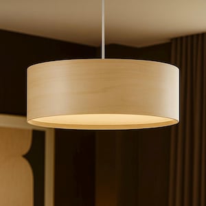 May include: A light-colored, cylindrical pendant lamp hangs from the ceiling. The lamp has a wooden shade and emits a warm, inviting glow. The design is simple and modern, suitable for various interior styles.