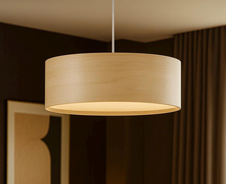May include: A light-colored, cylindrical pendant lamp hangs from the ceiling. The lamp has a wooden shade and emits a warm, inviting glow. The design is simple and modern, suitable for various interior styles.