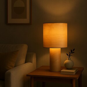 May include: A wooden table lamp with a cylindrical base and a lampshade. The lamp is on a wooden side table, next to a small vase with greenery and a book. A framed artwork hangs on the wall.