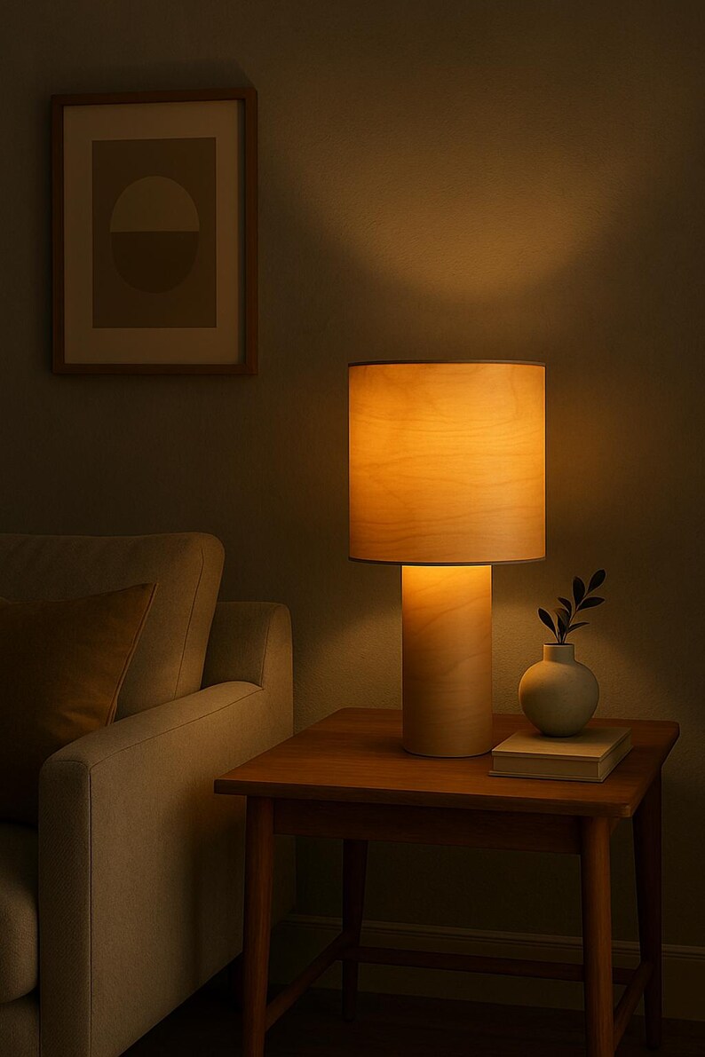 May include: A wooden table lamp with a cylindrical base and a lampshade. The lamp is on a wooden side table, next to a small vase with greenery and a book. A framed artwork hangs on the wall.