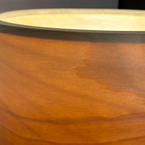 May include: A cylindrical wooden lamp with a warm, glowing interior. The lamp's exterior is a rich, reddish-brown with visible wood grain. The top edge is a darker brown, creating a contrasting border. The light source is not visible.
