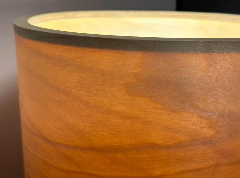 May include: A cylindrical wooden lamp with a warm, glowing interior. The lamp's exterior is a rich, reddish-brown with visible wood grain. The top edge is a darker brown, creating a contrasting border. The light source is not visible.
