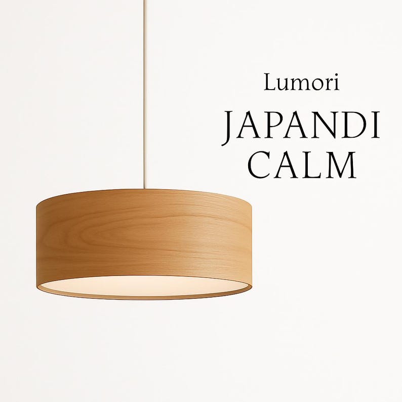 May include: A cylindrical pendant light fixture made of light-colored wood, suspended from a thin white cord. The fixture has a warm, inviting glow from the illuminated interior. The text "Lumori JAPANDI CALM" is displayed on the right.