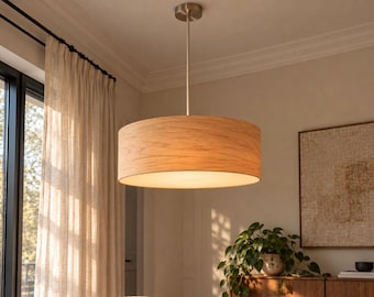 May include: A cylindrical pendant light fixture featuring a light-toned wood veneer drum shade. The lamp is suspended from the ceiling by a thin metal rod and a circular canopy, emitting a warm, diffused glow from the bottom opening.