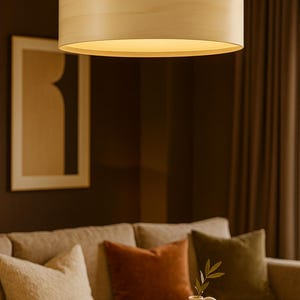 May include: A cylindrical wooden pendant light fixture hangs from the ceiling, illuminating a living room. The light fixture is made of light-colored wood. The room features a beige sofa with decorative pillows, a vase, and a candle.