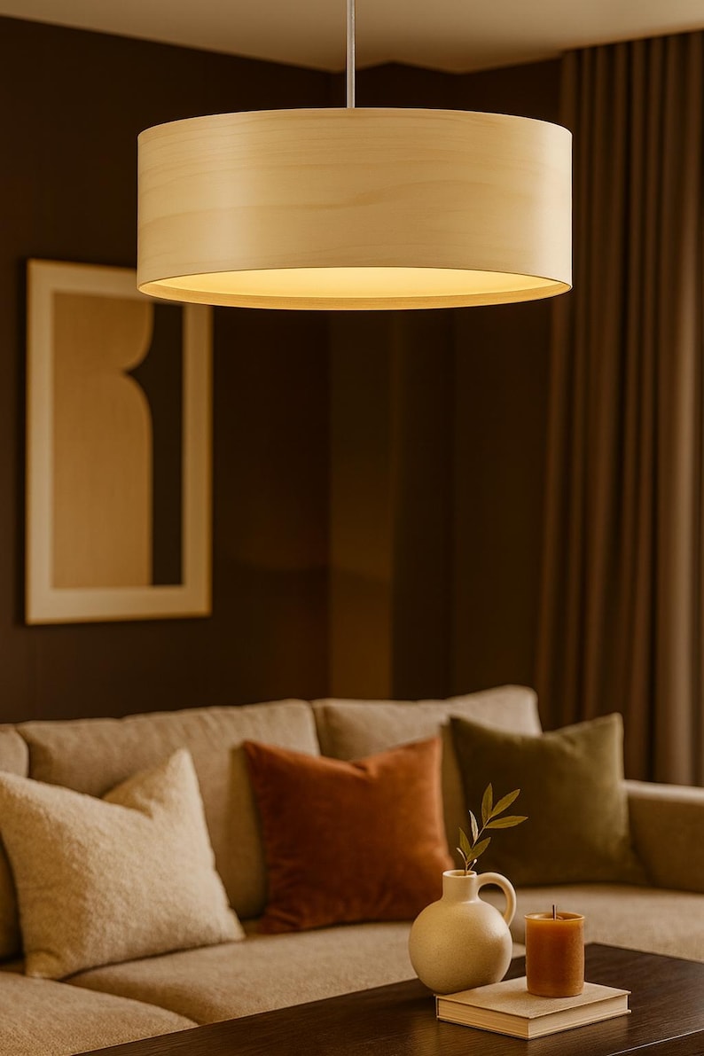 May include: A cylindrical wooden pendant light fixture hangs from the ceiling, illuminating a living room. The light fixture is made of light-colored wood. The room features a beige sofa with decorative pillows, a vase, and a candle.
