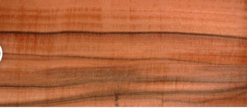 May include: Close-up of a piece of wood with a reddish-brown hue. The wood grain features dark, wavy lines that run horizontally across the surface. The texture appears smooth, with visible variations in color and pattern.