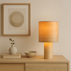 May include: A wooden table lamp with a cylindrical base and lampshade. The lampshade is a light wood color, and the base is a similar tone. A framed artwork, a small vase, a bowl, and a stack of books are also on the table.