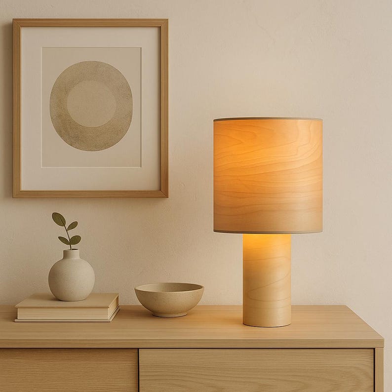 May include: A wooden table lamp with a cylindrical base and lampshade. The lampshade is a light wood color, and the base is a similar tone. A framed artwork, a small vase, a bowl, and a stack of books are also on the table.