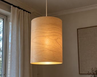 May include: A cylindrical pendant light fixture featuring a light-toned wood veneer shade with a visible natural grain pattern. The lamp is suspended from the ceiling by a thin, straight rod attached to a circular metallic canopy in a silver finish.