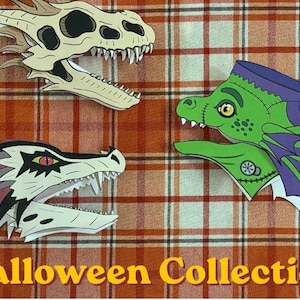 Peut inclure: Trois découpages en papier de têtes de dragon, dont un crâne beige, une créature verte avec des ailes violettes et des points de suture, et un dragon blanc avec des marques rouges et noires. Le texte Halloween Collection est imprimé en jaune.