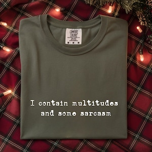 May include: Olive green t-shirt with the text "I contain multitudes and some sarcasm" in white. The shirt is folded and has a Comfort Colors tag. The background is a red and black plaid pattern.
