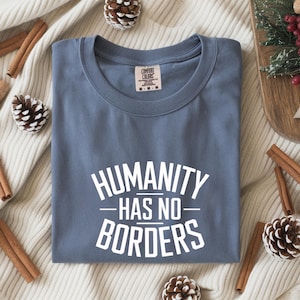 May include: A dusty blue t-shirt with the text "HUMANITY HAS NO BORDERS" in white. The shirt is folded neatly on a cream-colored surface, surrounded by cinnamon sticks, pine cones, and a wooden tray with greenery.