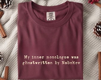 Puede incluir: Una camiseta Comfort Colors color granate con el texto "My inner monologue was ghostwritten by Nabokov" en fuente serif. La camiseta está doblada y expuesta sobre una superficie neutra con piñas y palitos de canela.