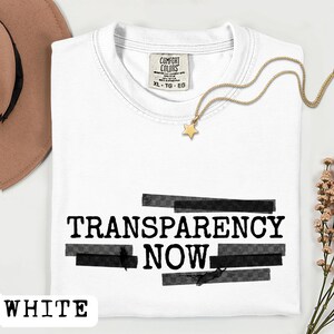 May include: White t-shirt with the words "TRANSPARENCY NOW" in black block letters, overlaid with gray rectangles. A gold star pendant necklace and a brown hat are also visible. The shirt has a "Comfort Colors" tag.
