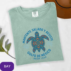 May include: A light green Comfort Colors t-shirt with a round neck. The shirt features a colorful turtle design with the text "MANTENTE SALADO Y RESISTE" and "GOLFO DE MÉXICO DESDE 1550". A brown hat is partially visible in the background.
