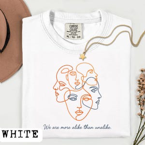 May include: White t-shirt with an orange and blue line art design of multiple faces. The text "We are more alike than unalike" is below the design. A gold necklace with a star pendant is on the shirt.