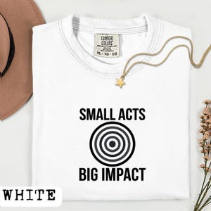 May include: White t-shirt with the text "SMALL ACTS BIG IMPACT" in black. The design includes a target graphic. A gold necklace with a star pendant rests on the shirt. The shirt has a Comfort Colors tag.