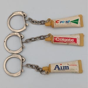 May include: Three vintage toothpaste tube keychains. The keychains feature miniature tubes of Crest, Colgate, and Aim toothpastes. Each tube is attached to a silver-colored keyring with a chain.