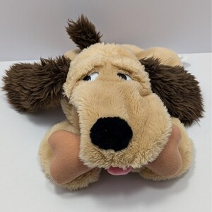 May include: A plush dog toy with a tan body, brown ears, and a black nose. The dog is holding a tan bone in its mouth and has a pink tongue sticking out. The toy has a sad expression.