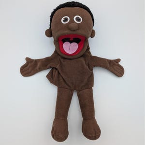 Può includere: Un burattino marrone con una grande bocca aperta, capelli neri e grandi occhi bordati di bianco. La bocca del burattino è foderata di rosso e ha una lingua rosa. Il burattino ha le braccia tese ed è progettato per essere indossato sulla mano.