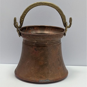 May include: A small, antique copper bucket with a brass handle. The bucket has a flared base and a cylindrical body with a decorative band near the top. The handle is arched and attached to the bucket with ornate brackets.