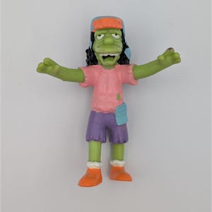 May include: A figurine of a green-skinned character with black hair, wearing a pink shirt, purple shorts, and orange boots. The character has a blue and orange hat and is posed with arms outstretched.