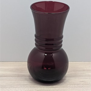 Può includere: Un vaso di vetro bordeaux scuro con base bulbosa e parte superiore svasata. Il vaso presenta una serie di scanalature orizzontali attorno al collo. Il vetro è liscio e riflettente, con una sfumatura da scuro a chiaro.