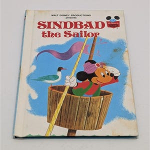 May include: Vintage Walt Disney Productions book titled "Sindbad the Sailor." The cover features Mickey Mouse dressed as a sailor in a wooden barrel, with a seagull and a mast with a purple flag against a blue sky background. The book has a white cover with red and black text.