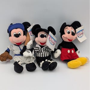 Könnte beinhalten: Drei Mickey Mouse Plüschtiere in Baseball-, Schiedsrichter- und klassischen Outfits. Der Baseball-Mickey trägt eine blaue Kappe und eine gestreifte Uniform und hält einen Schläger. Der Schiedsrichter-Mickey trägt ein schwarz-weiß gestreiftes Hemd. Der klassische Mickey hat rote Shorts.