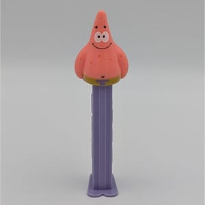 May include: A pink Patrick Star PEZ dispenser. The dispenser features a smiling Patrick Star character head with black eyes and a yellow waistband. The dispenser's stem is a light purple color.