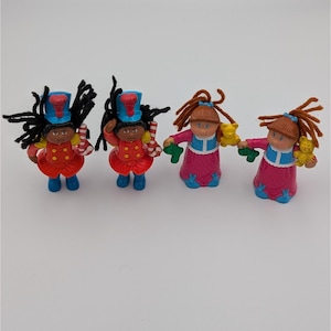 May include: Four small figurines. Two are dressed in red jackets and blue hats, holding candy canes. The other two wear pink dresses and hold small toys. All have painted faces and are standing on a white surface.