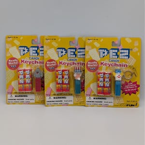 Può includere: Tre set di portachiavi PEZ Candy su confezione gialla. Ogni set include un dispenser PEZ e un portachiavi. I dispenser presentano personaggi come un clown, lo Zio Sam e una figura grigia. La confezione include il testo "PEZ Candy Keychain".