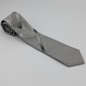 Puede incluir: Una corbata gris con un sutil patrón de rayas y líneas diagonales. La corbata está enrollada en un extremo, con el otro extremo extendiéndose hacia el espectador. El diseño de la corbata presenta una mezcla de texturas y tonos grises.