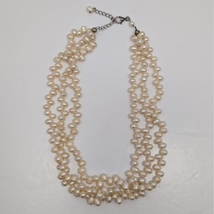 Könnte beinhalten: Eine mehrsträngige Perlenkette mit einer silberfarbenen Kette und einem Verschluss. Die Halskette besteht aus mehreren Strängen aus cremefarbenen, oval geformten Perlen. Die Perlen sind zu einem mehrschichtigen Effekt aufgereiht, geeignet für formelle oder informelle Anlässe.