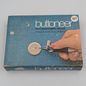 May include: A vintage "Buttoneer" box in blue with the text "Buttoneer" and "the 5-second button attacher." The box features an illustration of a hand using the button attacher to attach a button. The price tag shows $4.99.