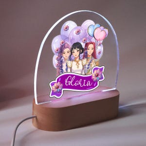 Op de afbeelding: Decoratieve LED-nachtlamp met een houten voet en een helder acryl paneel. Het paneel toont een cartoon illustratie van drie vrouwen, ballonnen en de naam "Gloria" op een paarse banner.