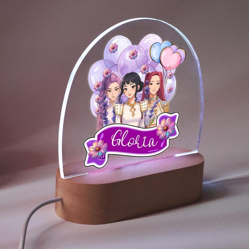 Op de afbeelding: Decoratieve LED-nachtlamp met een houten voet en een helder acryl paneel. Het paneel toont een cartoon illustratie van drie vrouwen, ballonnen en de naam "Gloria" op een paarse banner.