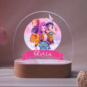 Op de afbeelding: Een gepersonaliseerd nachtlampje met een anime-achtige illustratie van drie vrouwelijke personages en een blauwe kat. De naam "Gloria" staat op een roze banner. Het licht staat op een houten voet.