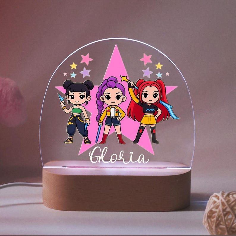 Op de afbeelding: Een gepersonaliseerd nachtlampje met drie stripfiguren in kleurrijke outfits en accessoires, tegen een roze ster en een helder acryl paneel. De naam "Gloria" staat eronder in cursief. De basis is van hout.