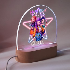 Op de afbeelding: Decoratief LED-nachtlampje met een cartoonillustratie van drie vrouwen en een blauwe kat. Het ontwerp is geplaatst tegen een stervormig neonlicht, met de naam "Gloria" aan de basis. Het licht staat op een houten voet.