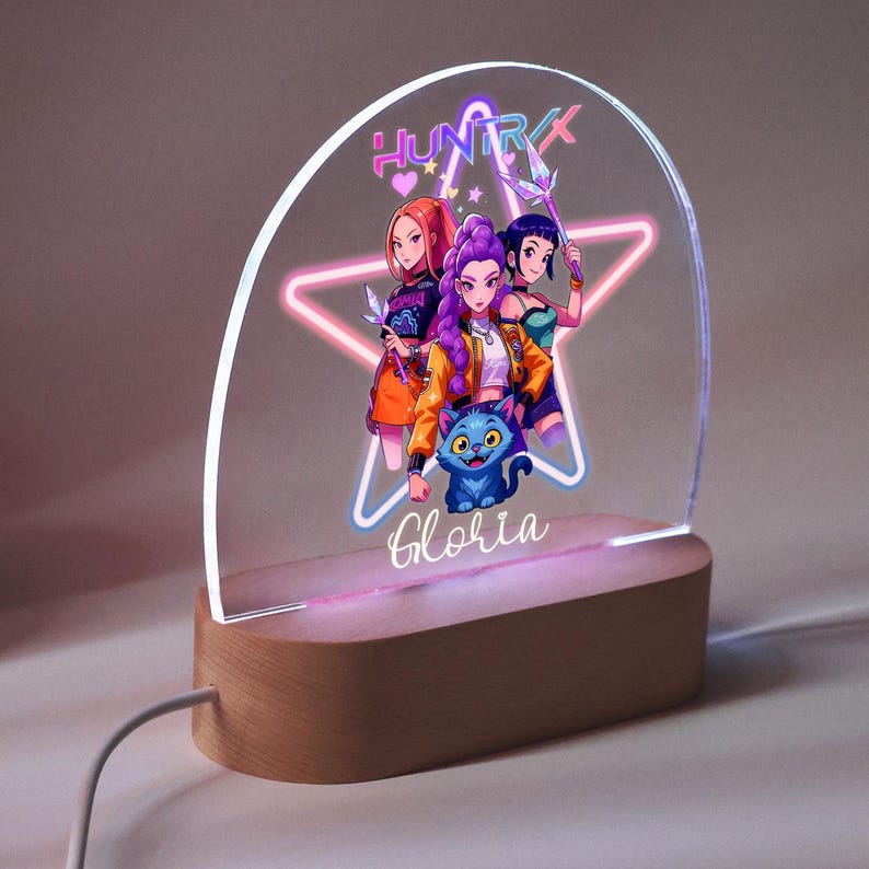 Op de afbeelding: Decoratief LED-nachtlampje met een cartoonillustratie van drie vrouwen en een blauwe kat. Het ontwerp is geplaatst tegen een stervormig neonlicht, met de naam "Gloria" aan de basis. Het licht staat op een houten voet.