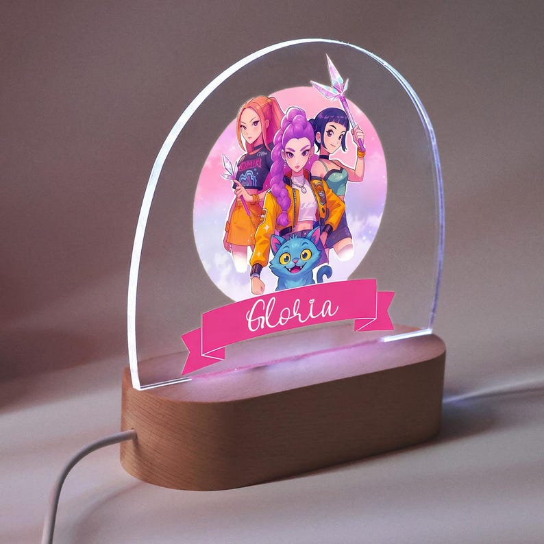 Op de afbeelding: Decoratieve LED-lamp met een kleurrijke anime-stijl illustratie van drie figuren en een blauwe kat. De lamp heeft een houten voet en een helder, gebogen acryl paneel. De naam "Gloria" staat op een roze banner.