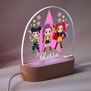 Op de afbeelding: Een decoratief nachtlampje met drie stripfiguren in kleurrijke outfits, tegen een roze ster en steraccenten. Het licht is gemonteerd op een houten voet, met de naam "Gloria" in cursief geschreven.
