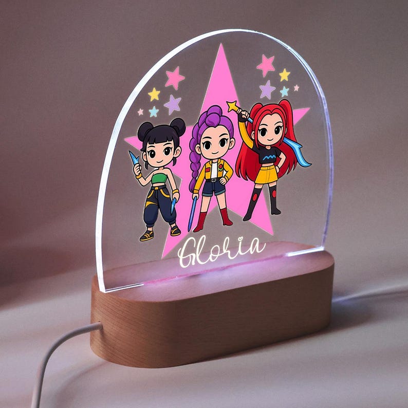 Op de afbeelding: Een decoratief nachtlampje met drie stripfiguren in kleurrijke outfits, tegen een roze ster en steraccenten. Het licht is gemonteerd op een houten voet, met de naam "Gloria" in cursief geschreven.