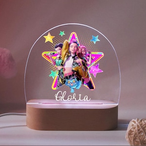 Puede incluir: Luz de noche decorativa con base de madera y parte superior de acrílico transparente. La luz presenta un gráfico de tres mujeres frente a una estrella, con el nombre "Gloria" debajo. El diseño crea un brillo ambiental cálido.
