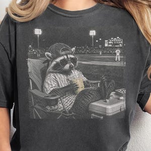 Può includere: Una t-shirt a maniche corte grigio antracite con una stampa grafica di un procione che indossa un berretto da baseball e occhiali da sole, seduto su una sedia pieghevole in uno stadio di baseball mentre tiene un bicchiere di plastica con cannuccia.