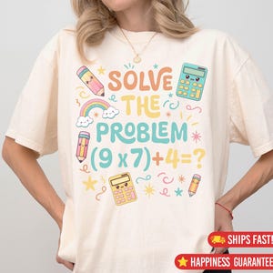 Può includere: T-shirt color crema con una grafica colorata che dice "SOLVE THE PROBLEM (9 x 7) + 4 = ?". Il design include matite dei cartoni animati, calcolatrici, un arcobaleno e stelle. Una maglietta divertente ed educativa.